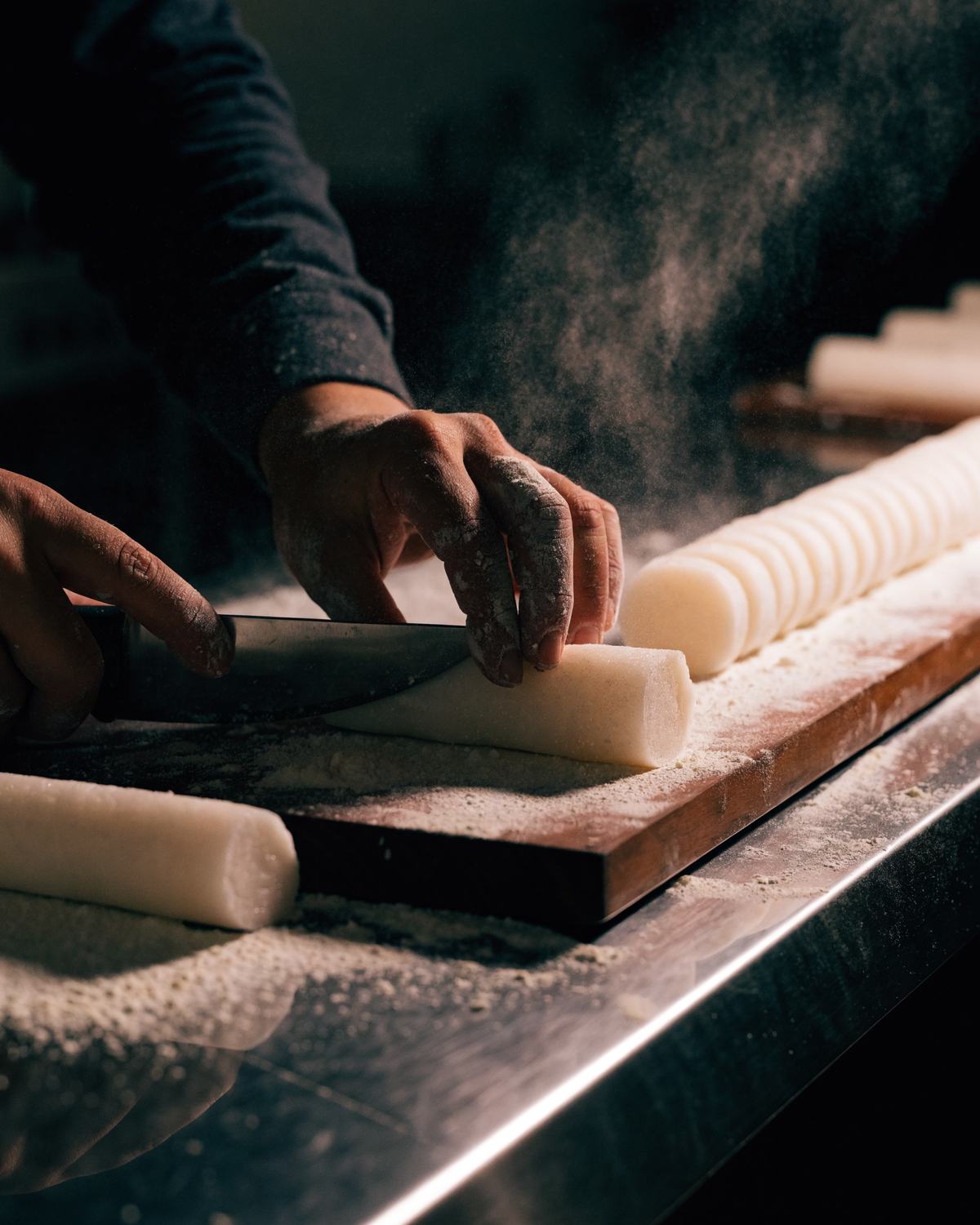 Hands slicing fresh rice cake logs on a floured board
